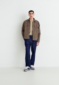 Brown button-up overshirt, beige t-shirt, blue denim pants, and white sneakers with blue accents. Casual layered outfit on a model.