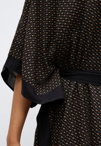 Robe en tissu noir avec un motif géométrique beige, manches courtes et taille marquée. Le matériau semble doux avec une légère brillance.