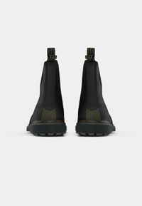 Uma par de botas pretas de tornozelo com abas para puxar, solas grossas e painéis laterais elásticos, mostradas de costas sobre um fundo branco.