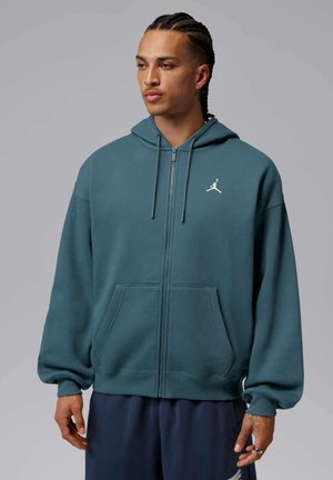Homme aux cheveux tressés porte un sweat à capuche zippé turquoise avec un logo blanc sur la poitrine gauche, debout devant un fond gris uni.