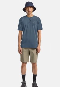 Camicia blu navy a maniche corte con un logo discreto, abbinata a pantaloncini color cammello fino al ginocchio, calzini neri e un cappello a secchiello blu.