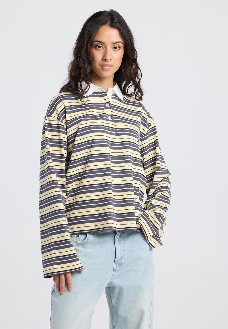 Nuori nainen, jolla on pitkät tummat hiukset, pukeutuneena löysään kelta-harmaa-valko-raitaiseen pitkähihaiseen paitaan ja vaaleansinisiin farkkuihin, seisoo valkoista taustaa vasten.