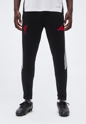 Henkilö, joka käyttää mustia Liverpool FC Adidas -treenihousuja valkoisilla raidoilla ja mustia jalkapallokenkiä valkoisella taustalla.