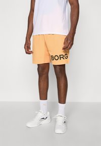 Orange shorts with black "BORG" text on the side, made of lightweight fabric. Paired with a white shirt and white sneakers.