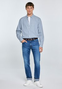 Camisa de cuadros azul clara, pantalones vaqueros azul claro, cinturón marrón, zapatillas blancas. Modelo de pie con una mano en la cadera, fondo blanco.