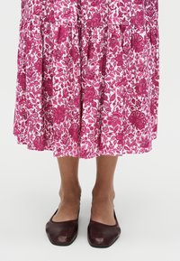 Κάτω μέρη ποδιών που φορούν ροζ midi φούστα με λουλούδια και σκούρα καφέ μπαλαρίνες με τετράγωνη μύτη, στέκονται σε απλό λευκό φόντο.