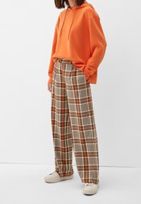Oranje hoodie met een kangoeroezak, gecombineerd met plooibijlen met brede pijpen in bruin en roomkleurige tinten. Witte sneakers maken de look compleet.