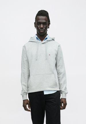 Mladý muž s tmavou pletí a zapletenými vlasy, který má na sobě světle šedou mikinu s kapucí přes modrou košili s límečkem a černé kalhoty, stojící před bílým pozadím.