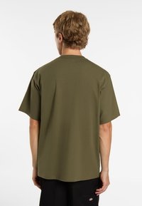 T-shirt in cotone verde oliva con maniche corte, scollo rotondo, taglio rilassato e orlo dritto. La vista posteriore mostra dettagli minimi e una consistenza morbida.