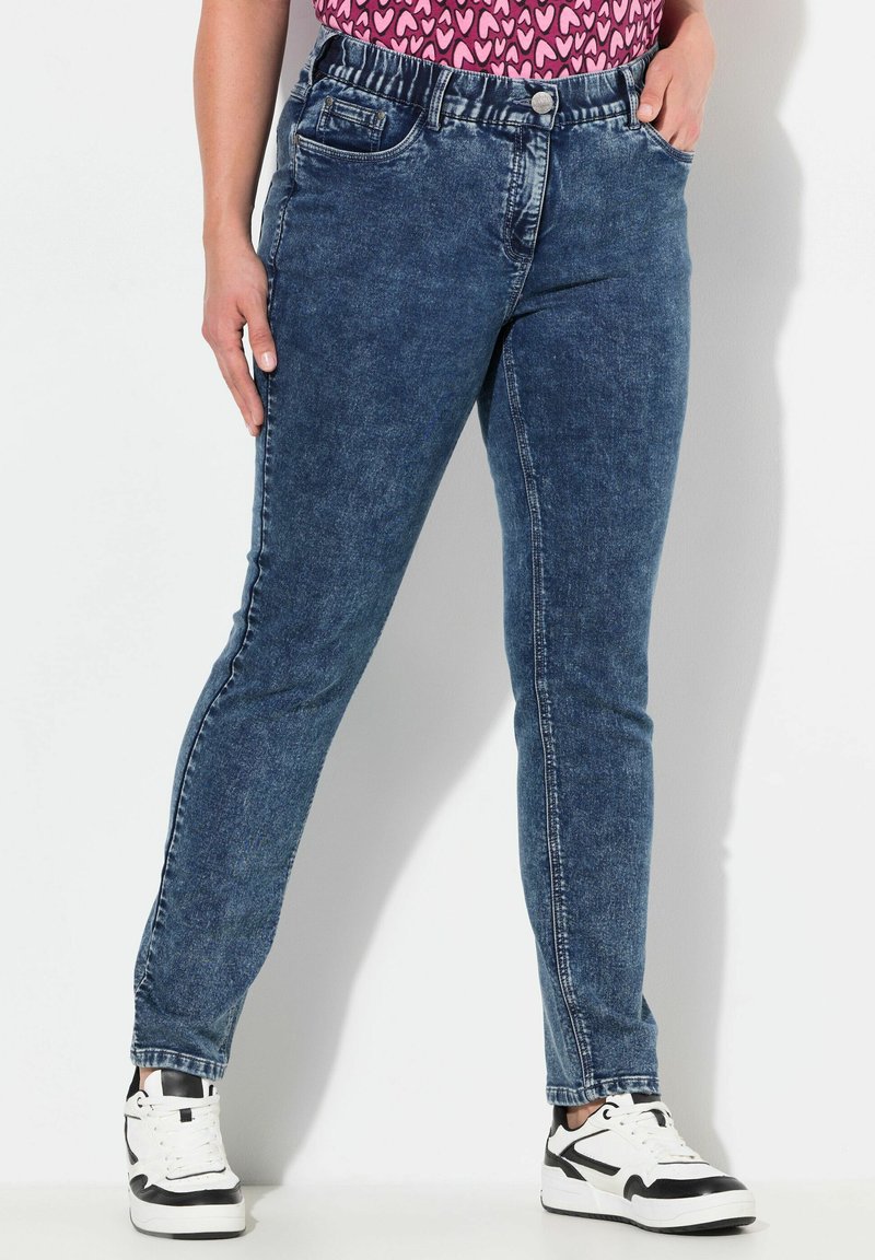 Jeans de mezclilla en azul oscuro con un corte ajustado, cintura elástica, bolsillos frontales y cierre de botón, combinados con zapatillas negras y blancas.