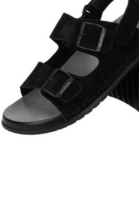 Schwarze offene Sandale aus Wildleder mit zwei verstellbaren Schnallen mit der Aufschrift "wojas" und einer dicken, profilierten Sohle.