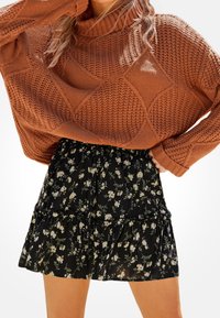 Hnědý pletený rolák s texturovaným diamantovým vzorem v kombinaci s černou květinovou mini sukní s volány, která má bílé květy.