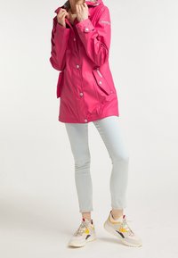 Giacca da pioggia impermeabile rosa con bottoni a pressione, cappuccio e tasche laterali, abbinata a jeans azzurri chiari e sneakers beige.
