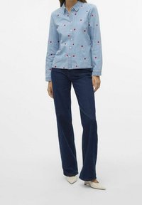 Camicia di jeans azzurra con ricamo a cuore rosso, chiusura a bottoni frontale, maniche lunghe; abbinata a jeans svasati blu scuro e scarpe beige con tacco basso.