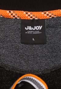 Zwarte en grijze stof met een gestructureerde afwerking, voorzien van een oranje gevlochten rand. Inclusief een label met "J&JOY" en maat "L."