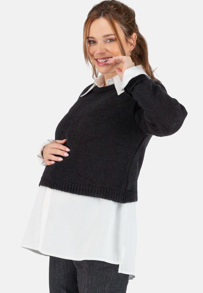 Schwarzer, gestrickter, kürzerer Pullover mit geripptem Saum über einem weißen Kragenhemd. Das Model posiert mit der Hand auf dem Bauch und trägt dunkle Hosen.