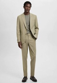 Lichtbeige pak met een getailleerde jas en slanke broeken, gecombineerd met een groene top. Gladde textuur en minimale details. Zwarte schoenen.