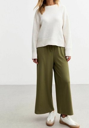 Žena v krémovém svetru, olivově zelených širokých kalhotách a bílých teniskách stojící na světle šedé podlaze před bílou zdí.