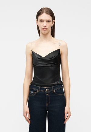 Γυναίκα που φοράει μαύρο σατέν τοπάκι με λουράκια από πέρλες και σκούρο μπλε τζιν ψηλόμεσο, στέκεται μπροστά σε ανοιχτό φόντο.