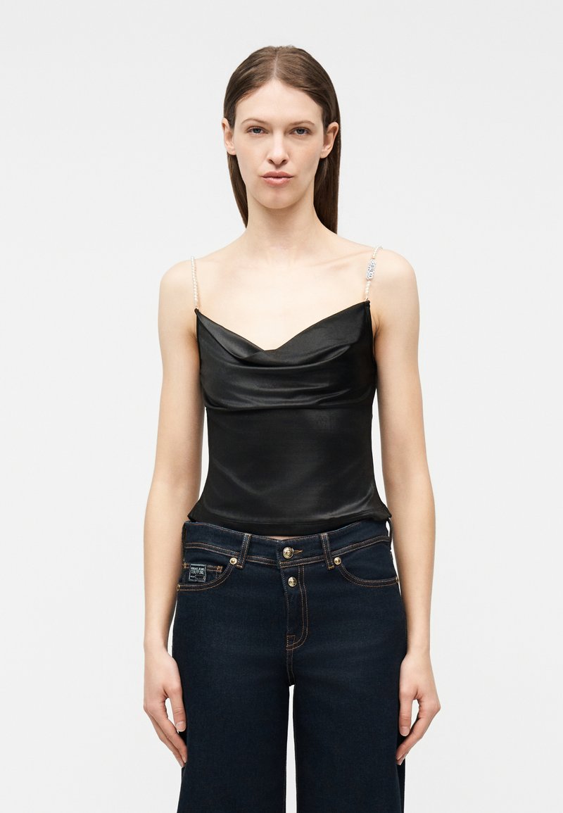 Γυναίκα που φοράει μαύρο σατέν τοπάκι με λουράκια από πέρλες και σκούρο μπλε τζιν ψηλόμεσο, στέκεται μπροστά σε ανοιχτό φόντο.