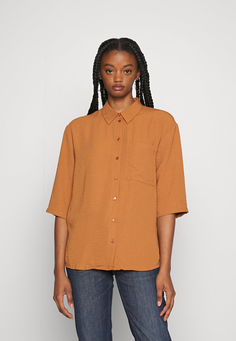 Camisa de botão de manga curta na cor laranja queimado, com tecido texturizado, um único bolso no peito e gola redonda, combinada com jeans de denim azul.