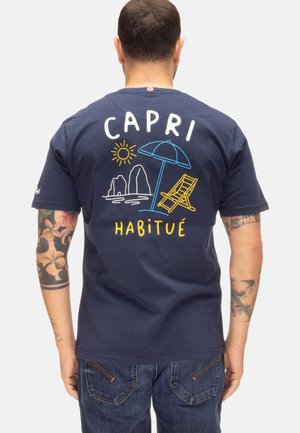 Uomo con braccia tatuate indossa una maglietta blu navy con un design colorato della spiaggia di Capri, che include sole, rocce, ombrellone e sdraio, e la scritta "Capri Habitué."
