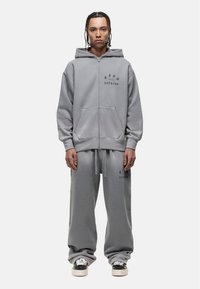 Gray zip-up hoodie with logo, paired with matching gray sweatpants. Material is soft, showcased in a relaxed fit. Worn with black and white sneakers.