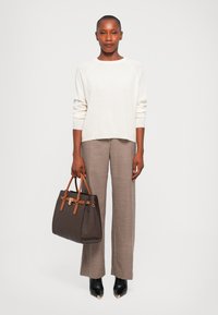 Femme portant un pull blanc et un pantalon pied-de-poule marron, tenant un sac à main marron, se tenant devant un fond clair.