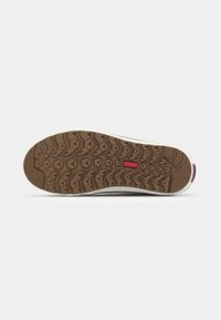 Semelle de chaussure avec une bande de roulement en caoutchouc marron présentant des motifs en chevrons et en forme de cœur, avec un petit logo rectangulaire rouge au centre.