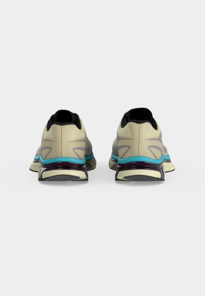 Ζευγάρι μπεζ παπούτσια τρεξίματος με μαύρη επένδυση, μπλε λεπτομέρειες στη μέση της σόλας και παχιές, διαφανείς μοβ υποστηρίξεις φτέρνας, που φαίνονται από πίσω.