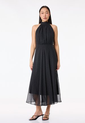 Kvinde iført sort ærmeløs halter midi kjole med hvide prikker og sorte sandaletter med hæl, stående mod en hvid baggrund.