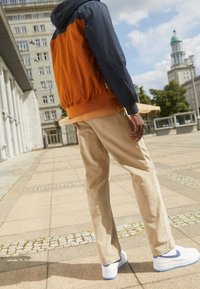 En tvåradig jacka med orange kropp och marinblå ärmar, i kombination med beiga kordbyxor och vita sneakers med blå detaljer.