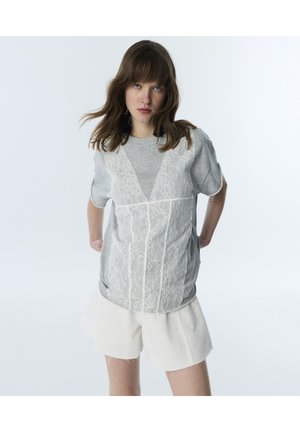Jonge vrouw met schouderlang bruin haar draagt een grijze top met kantoverlay en witte shorts, staand tegen een effen lichte achtergrond.