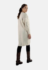 Lång, beige stickad kofta med texturerade ärmar. Bärs över svarta leggings och bruna knähöga stövlar, med en avslappnad passform och ribbade kanter.