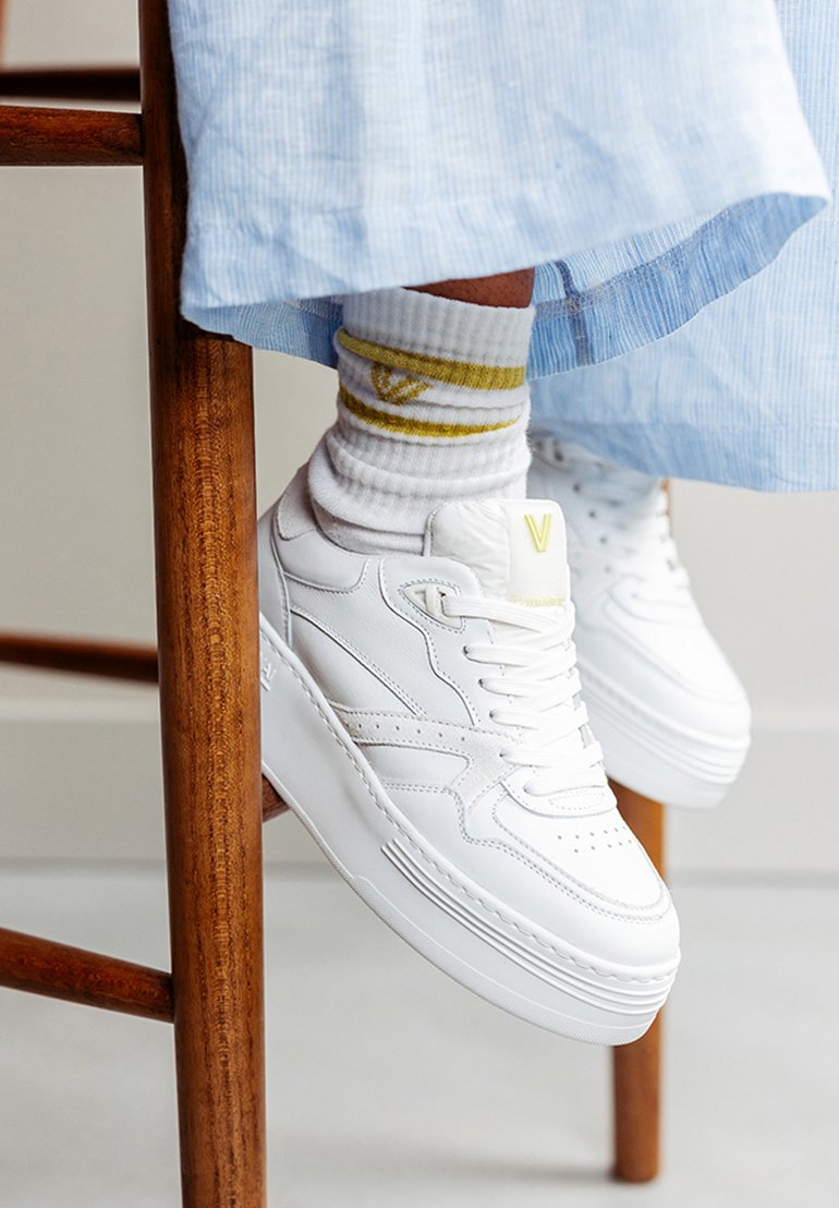 White leather sneakers with a chunky sole, featuring perforations and subtle branding. Paired with white socks that have yellow accents.