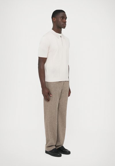 Πόλο μπλούζα σε ανοιχτό μπεζ πλεκτό με κοντά μανίκια και φερμουάρ μπροστά. Συνδυασμένη με υφασμένα, φαρδιά ανοιχτόκαφέ παντελόνια και μαύρα αθλητικά παπούτσια.