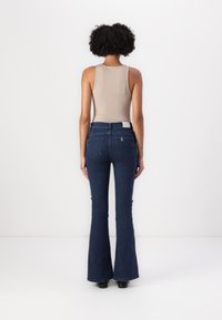 Jeans de denim acampanados en un lavado oscuro, diseño de cintura media con una cintura ajustada, detalles de costura sutiles y bolsillos traseros. Combinado con una camiseta sin mangas beige.