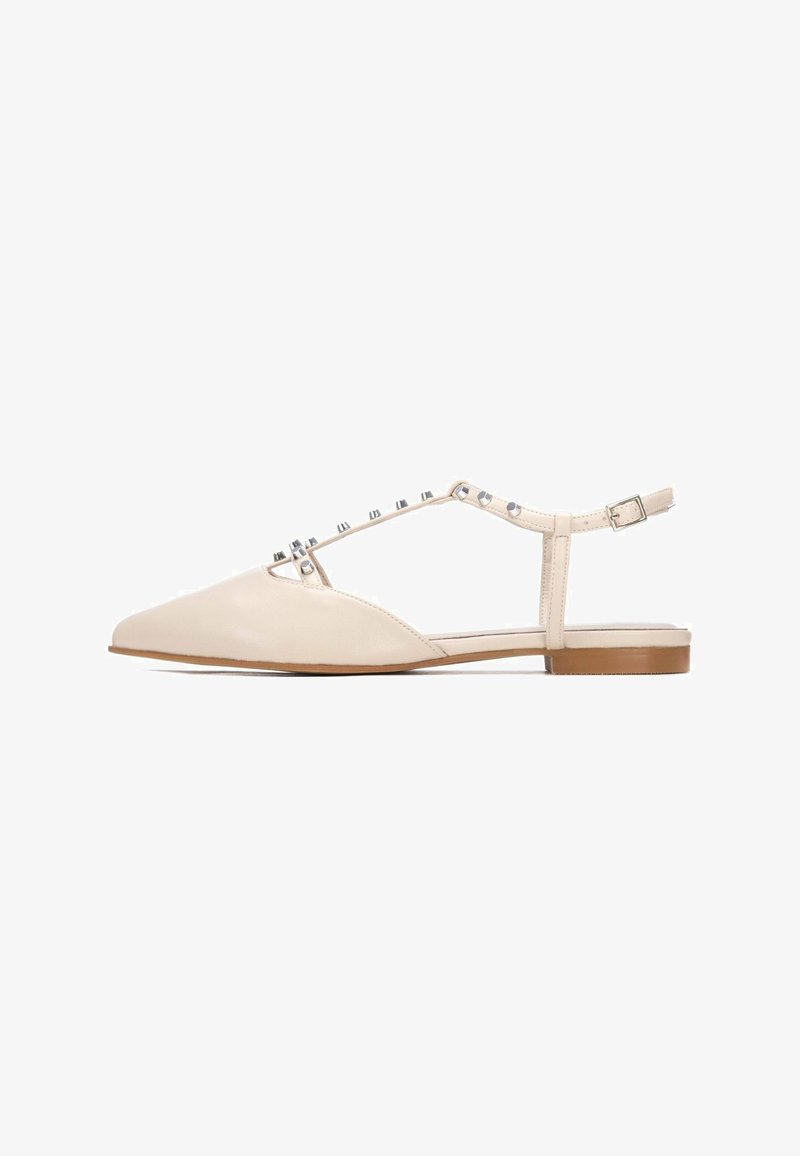 Zapatos planos de cuero beige con punta estrecha, respaldo abierto y correa ajustable en el tobillo con tachuelas metálicas decorativas en la parte frontal.