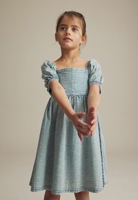 Hele sinine kleit lühikeste puhvärvadega, tekstureeritud kangast, ruudukujuline kaelus ja volditud seelik. Omab lihtsat äärist ja peent mustrit.