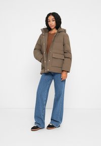 Brun dunjakke med hætte, der har trykknapper og to forlommer. Sammen med vide blå jeans og sort fodtøj.