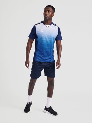 Athlète masculin debout, portant un maillot de sport à manches courtes bleu dégradé, un short bleu marine, des chaussettes blanches et des chaussures de sport noires sur un fond uni.