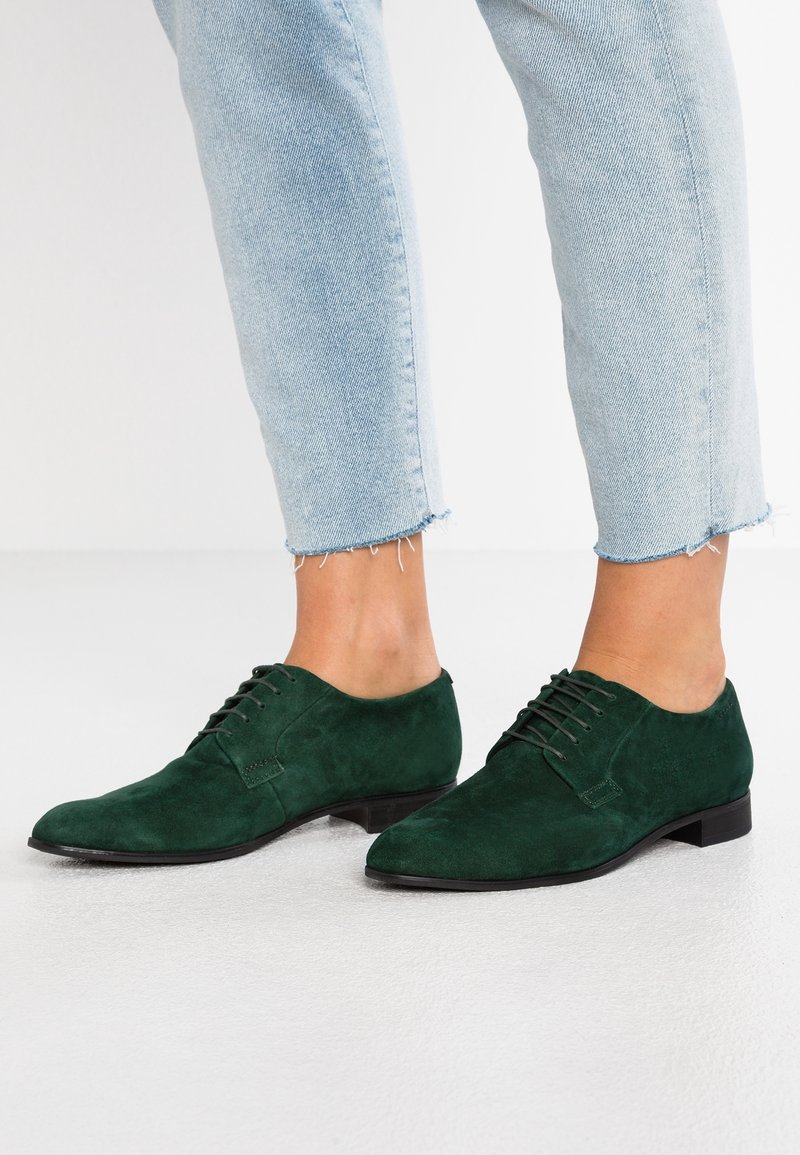 Personne portant un jean court bleu clair et des chaussures habillées à lacets en daim vert foncé sur un sol clair.