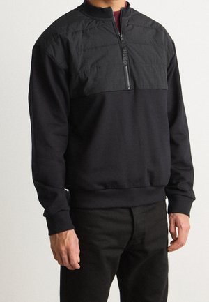 Hombre con pulóver negro con parte superior acolchada en el pecho y cierre de media cremallera, combinado con pantalones negros, sobre fondo liso.