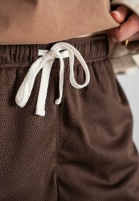 Pantaloni scurți din plasă maro, cu o suprafață texturată, talie elastică și șnur alb. Prezintă mici perforații pentru ventilare.