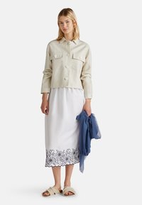 Helle beiger, kürzerer Mantel mit großen Knöpfen und zwei vorderen Taschen, über einem weißen Kleid mit floraler, marineblauer Stickerei am Saum. Beigefarbene Riemchensandalen und ein blauer Schal.