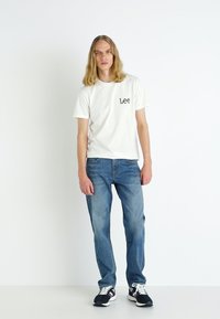 T-shirt branca de manga curta com o logótipo "Lee" bordado em preto, combinado com jeans de ganga azul. Modelo usa ténis pretos e brancos.