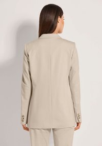 Beige op maat gemaakte blazer met een gladde textuur, enkele achterste split en knoopaccenten op de mouwen, gezien vanaf de achterkant.