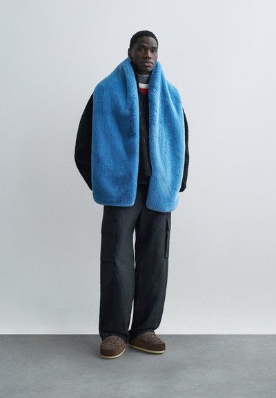 Μπλε περιτύλιγμα από ψεύτικη γούνα, μαύρο ανάγλυφο σακάκι, στρωμένο πουλόβερ, σκούρο παντελόνι cargo και καφέ κεντημένα παντόφλες δημιουργούν μια σύγχρονη εμφάνιση.