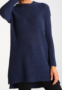 Robe-pull à manches longues texturée bleu marine portée avec des leggings noirs, modélisée avec une main touchant le cou et l'autre reposant le long du corps.