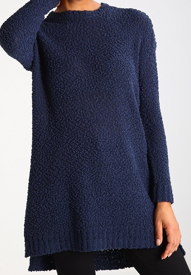 Robe-pull à manches longues texturée bleu marine portée avec des leggings noirs, modélisée avec une main touchant le cou et l'autre reposant le long du corps.